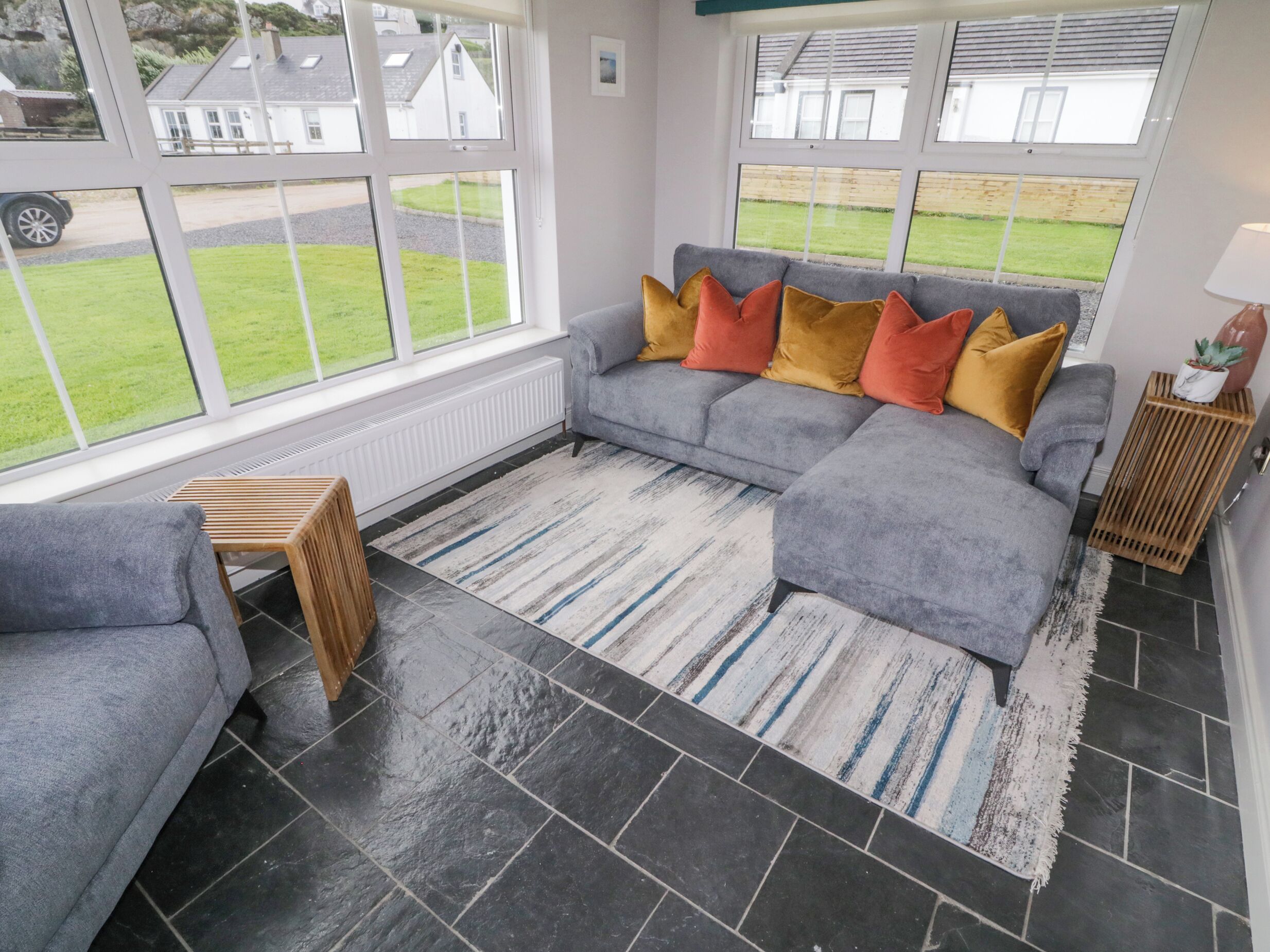 A cozy living room with a grey sofa, colorful cushions, a striped rug, and large windows offering a view of a green lawn and houses.
