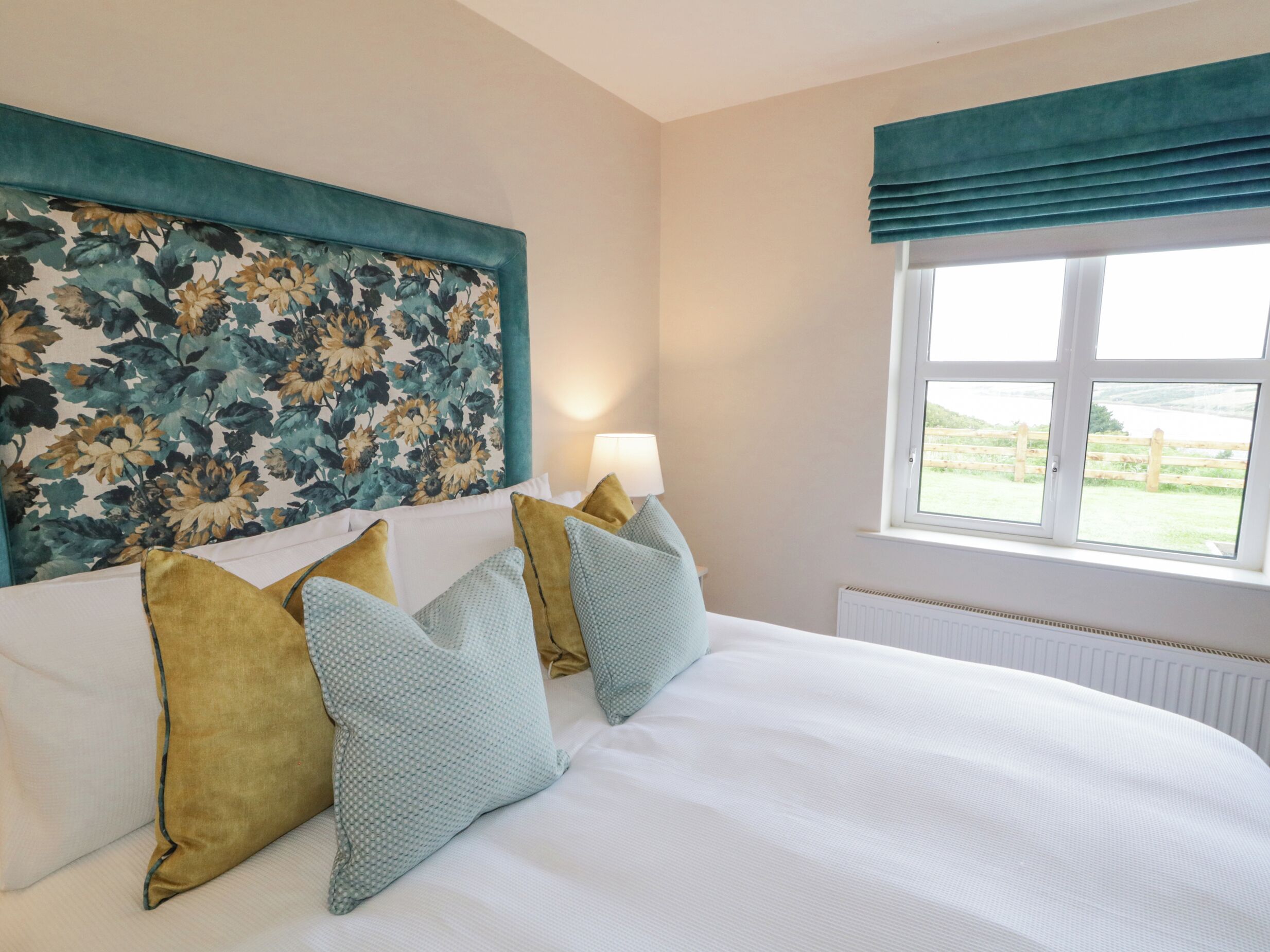 A neatly made bed with white linens and multiple pillows, a floral headboard, a window with a view of the outdoors, and a bedside lamp.