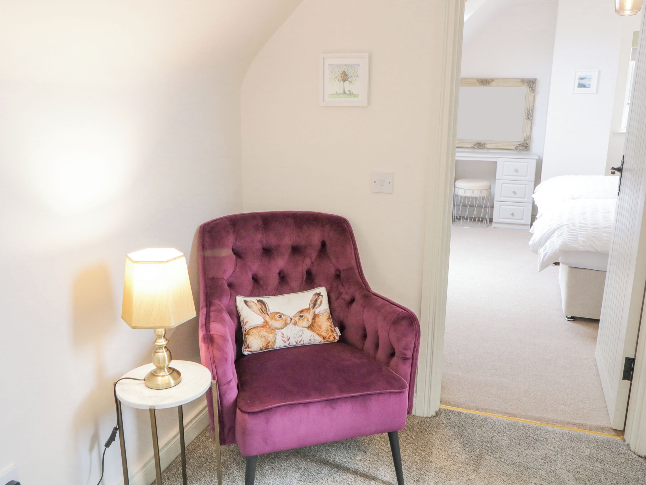 Een kamer met een paarse fluweelzitzak, een klein wit nachtkastje met een lamp, een ingelijst schilderij aan de muur, en een glimp van een slaapkamer met een bed en een dressoir.