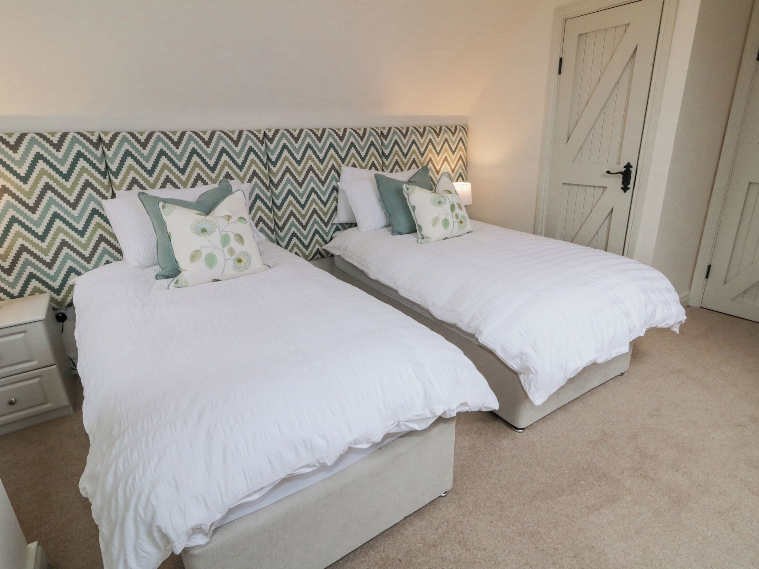 A double room with two beds, a patterned headboard, a white door, and a nightstand with a lamp.