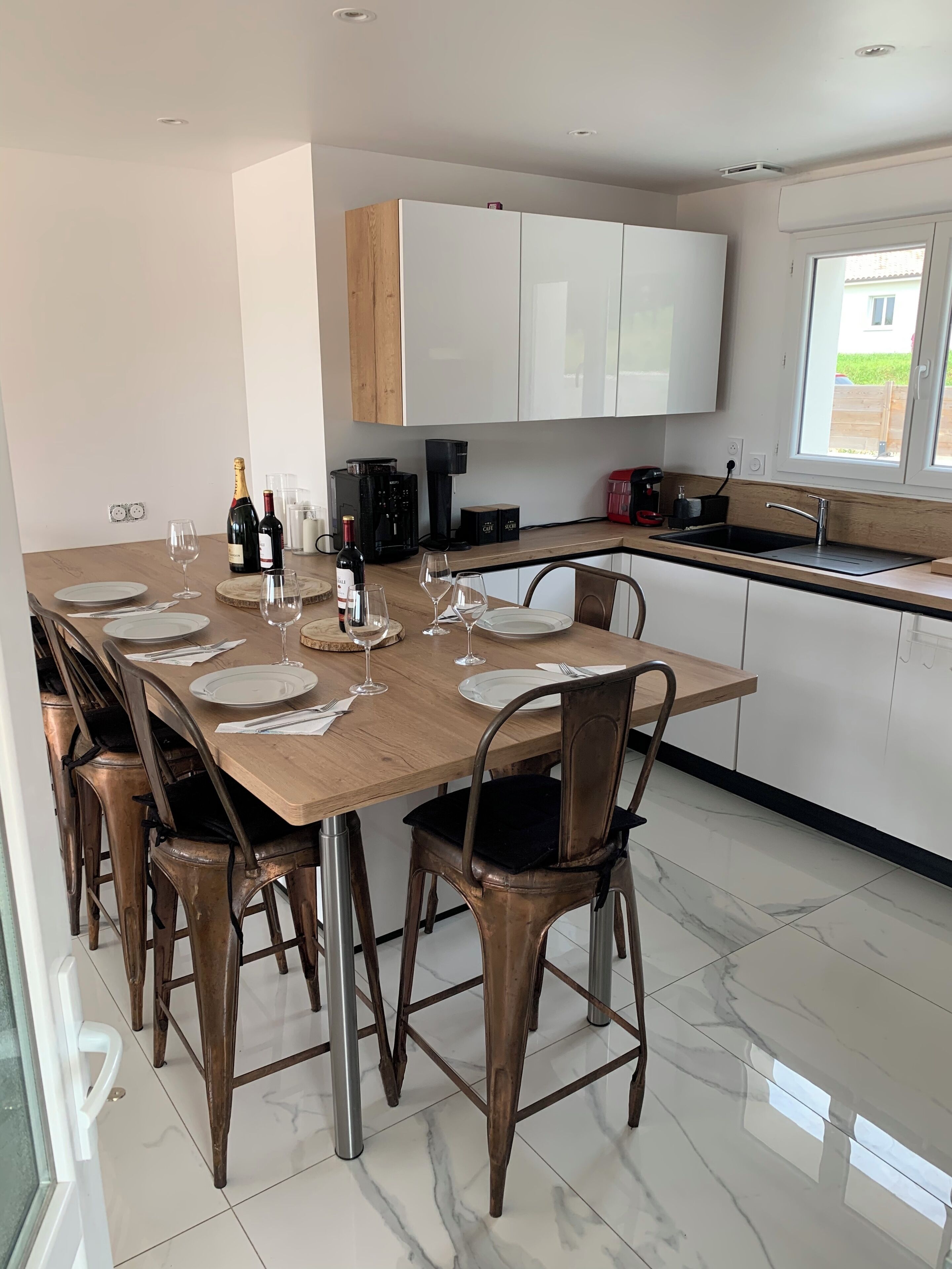 A modern kitchen with a large dining table set for six, equipped with wine glasses and bottles.