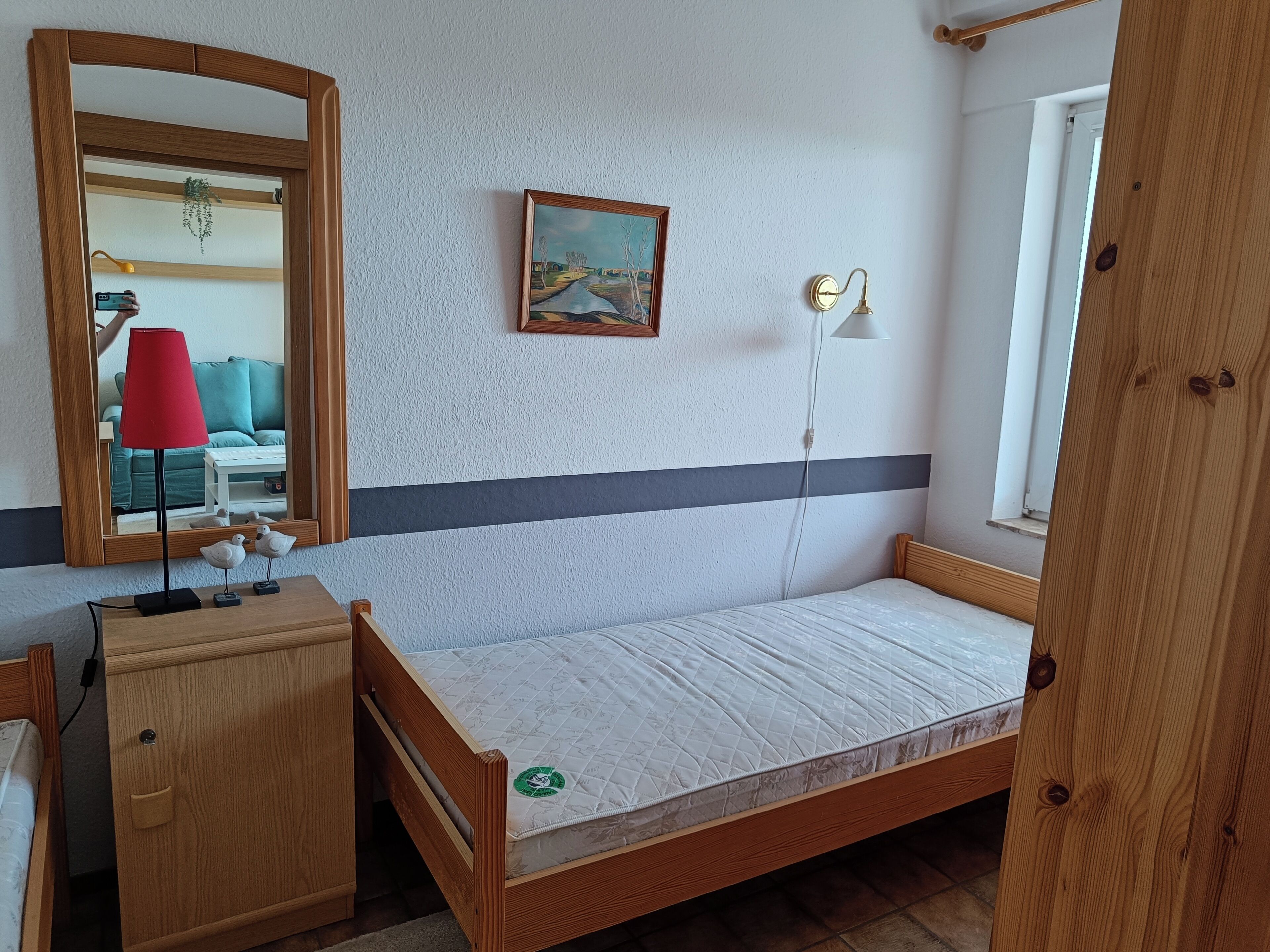 Ein kleiner, ordentlich eingerichteter Raum mit einem hölzernen Bett, einem Nachttisch aus Holz, einem Spiegel, einem Gemälde und einem Fenster.