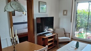 A cozy living room with a wooden dining table, white chairs, a TV, and a window with a view of greenery. - Ferienwohnung Sielluft (Dornum)
