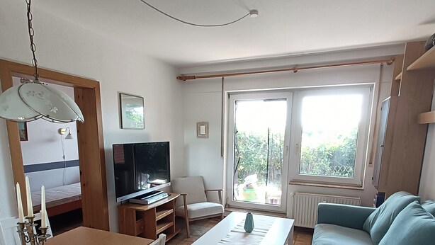 A living room with a sofa, a TV, a dining table with chairs, and a large window. - Ferienwohnung Sielluft (Dornum)