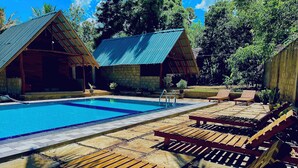 A pool area with wooden loungers, a clear blue pool, and two rustic cabins with metal roofs. - Maika safari (Udawalawa)