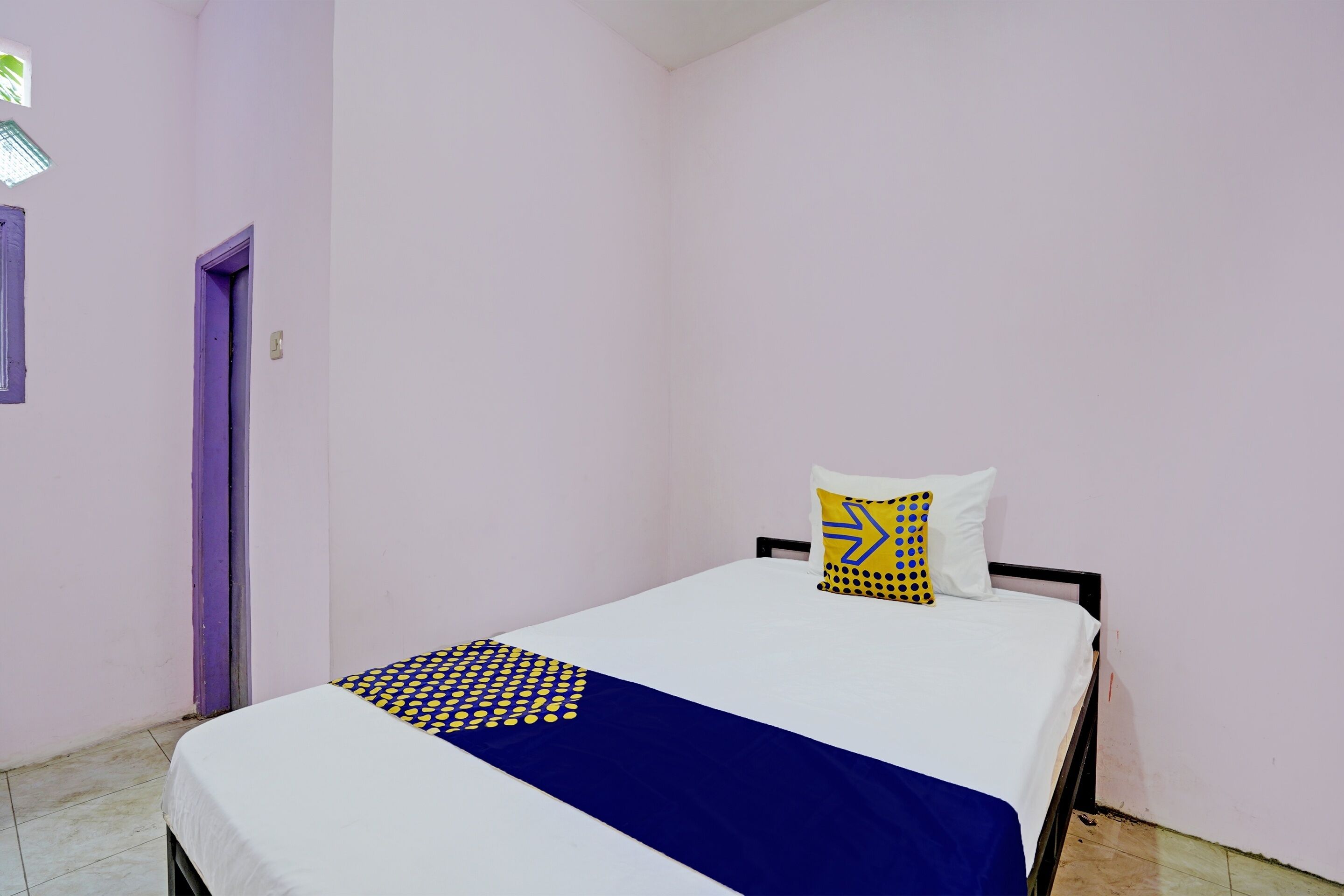 A single bed with a white and blue bedspread, a patterned pillow, and a purple door in a room with light-colored walls.