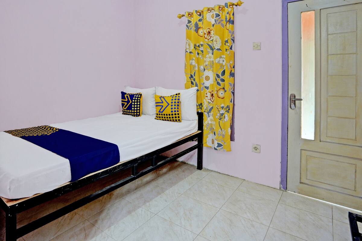 Una cama individual con una colcha azul y blanca, dos almohadas con diseños geométricos y una cortina floral amarilla.