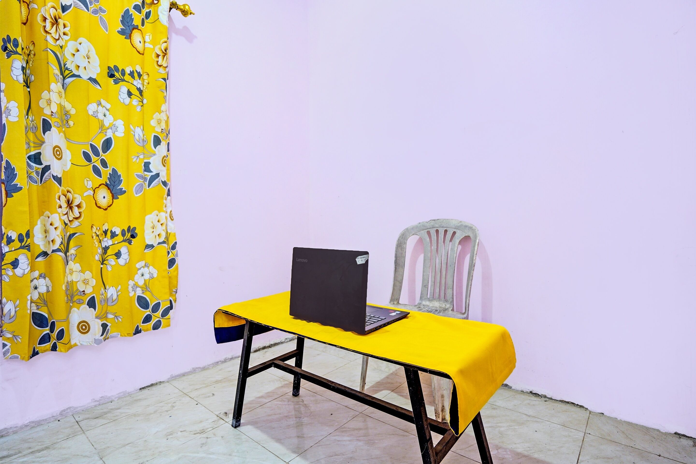 A yellow table with a laptop, a plastic chair, and a floral curtain in a room with a plain wall and tiled floor.