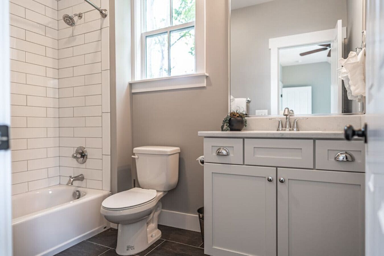Une salle de bain avec des toilettes blanches, une baignoire et une douche. Il y a une fenêtre au-dessus des toilettes, ainsi qu’un lavabo avec un miroir au-dessus du meuble-vasque.