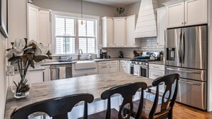 Una cocina moderna con muebles blancos, una isla grande con sillas, un refrigerador de acero inoxidable y una ventana que proporciona luz natural.