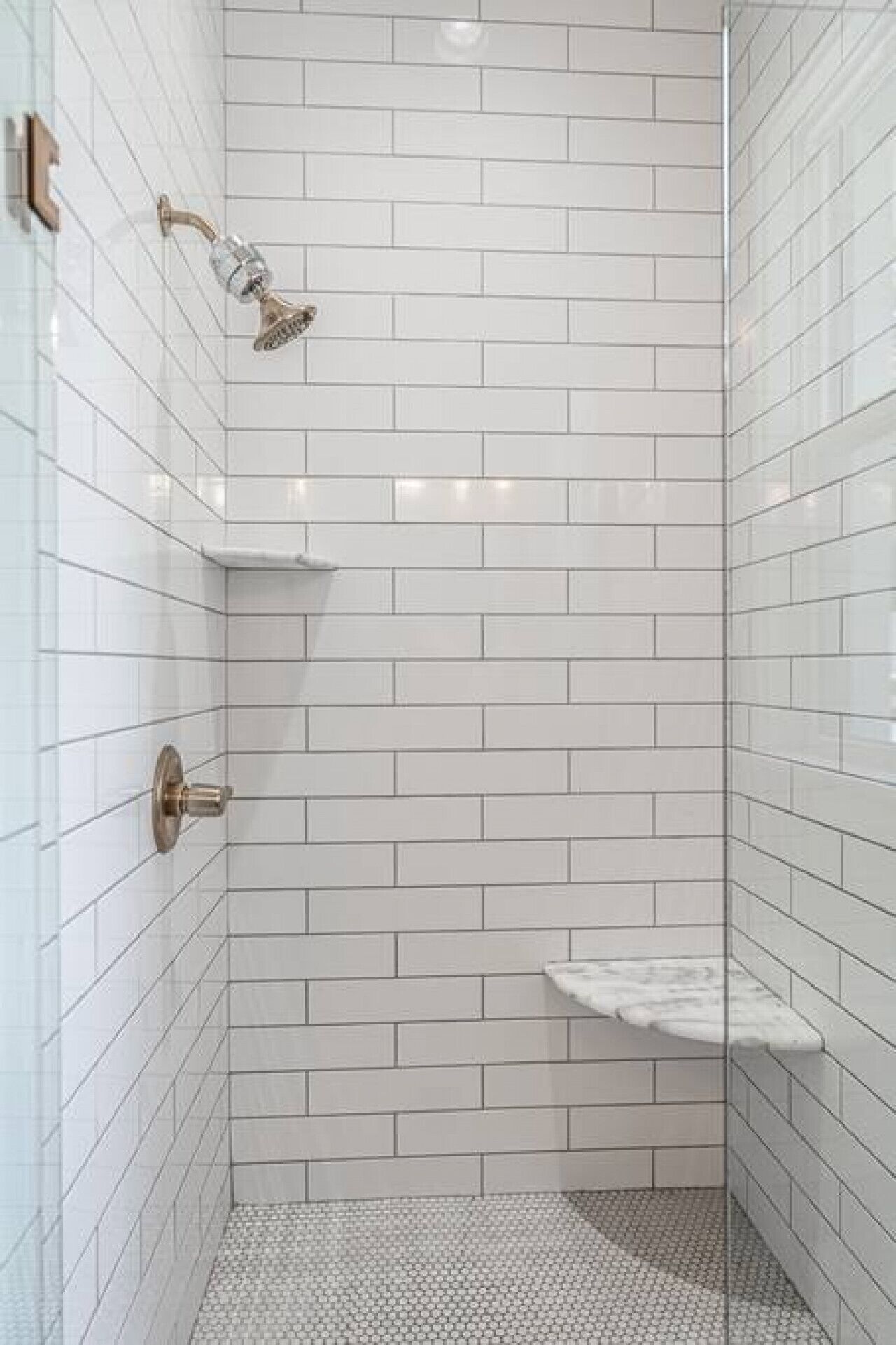A small, white-tiled shower stall with a glass shower head and a single shelf.