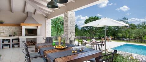 An outdoor patio with a long wooden table, chairs, a barbecue grill, and a pool in the background.
