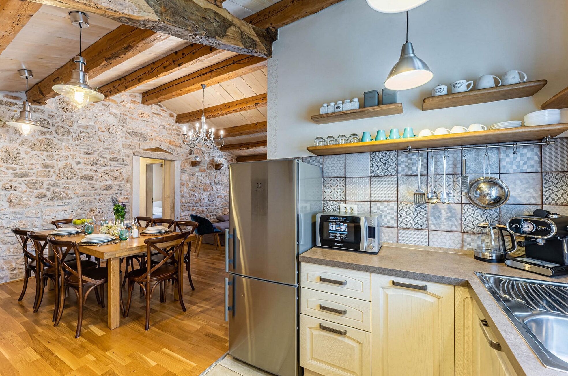 A kitchen with a stone wall, wooden ceiling beams, a stainless steel refrigerator, a microwave, and a coffee machine.