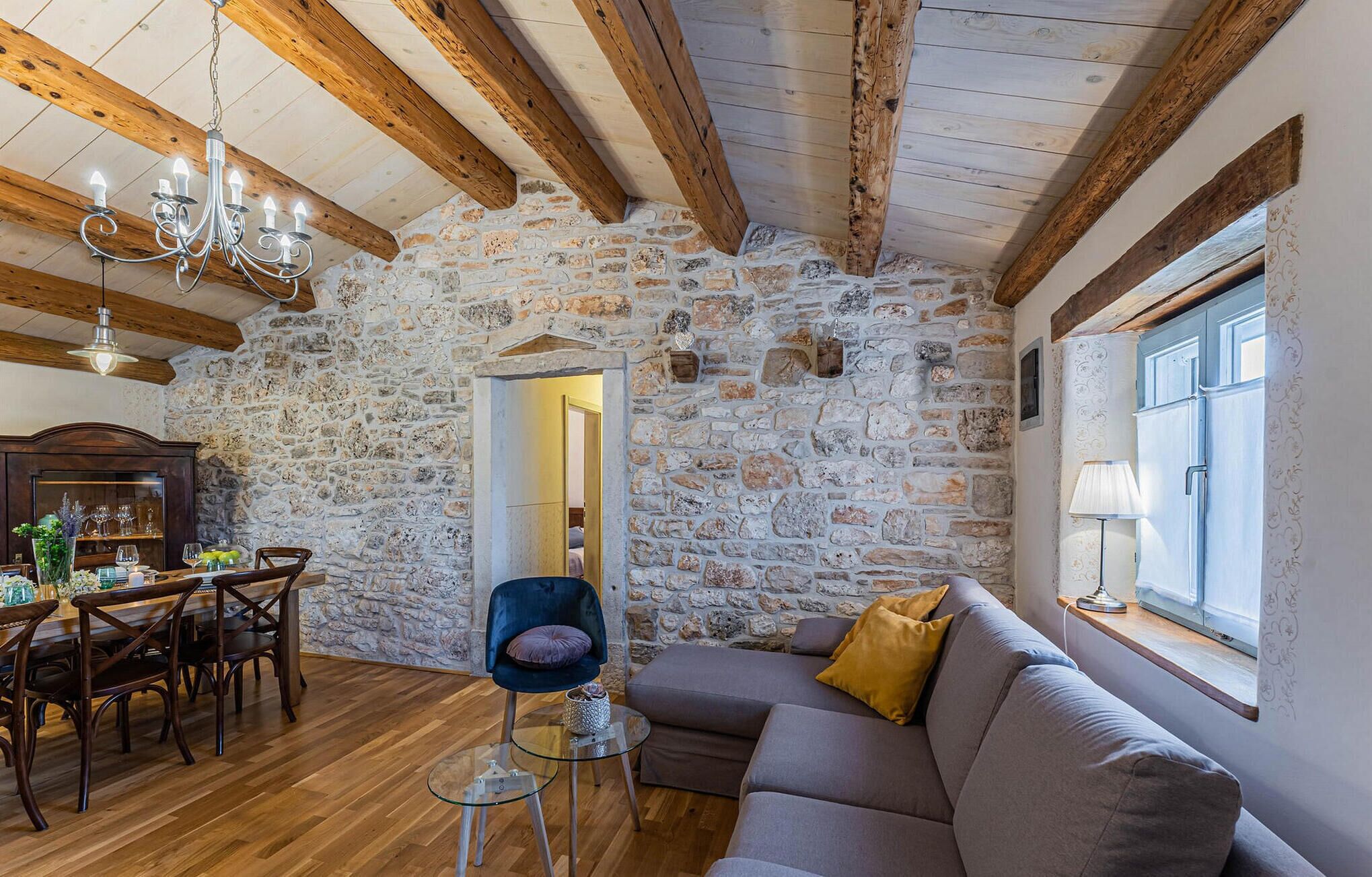 Een rustiek interieur met zichtbare houten balken, een stenen muur, een eethoek met een houten tafel en stoelen, en een moderne bank met uitzicht.