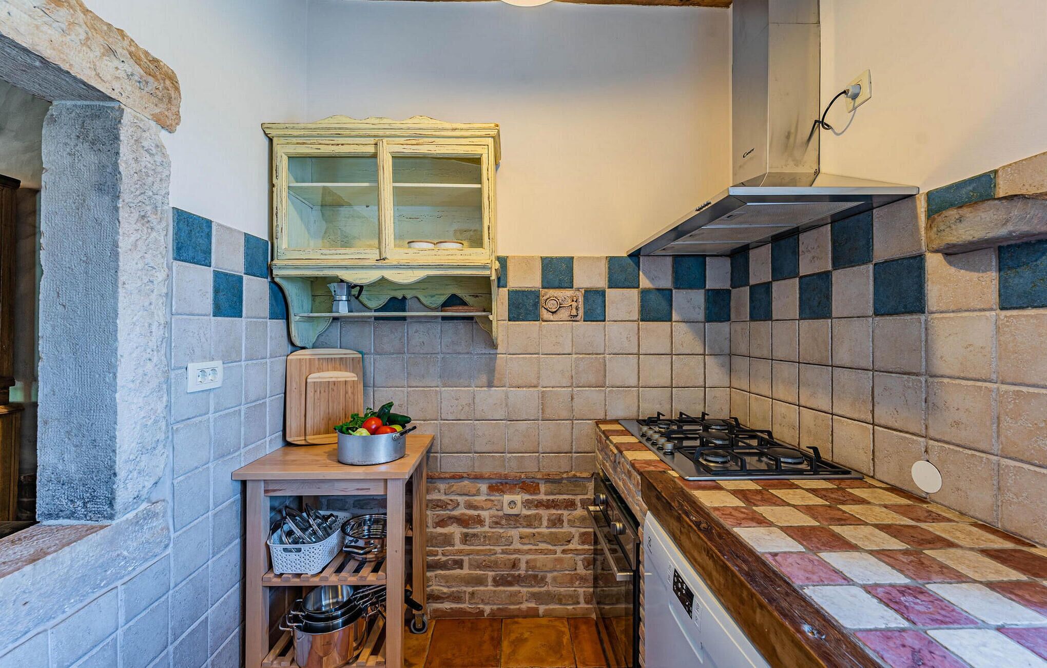 Une cuisine compacte équipée d’une cuisinière à gaz, d’une étagère en bois où sont rangés les ustensiles de cuisine, et d’une fenêtre donnant sur l’extérieur.