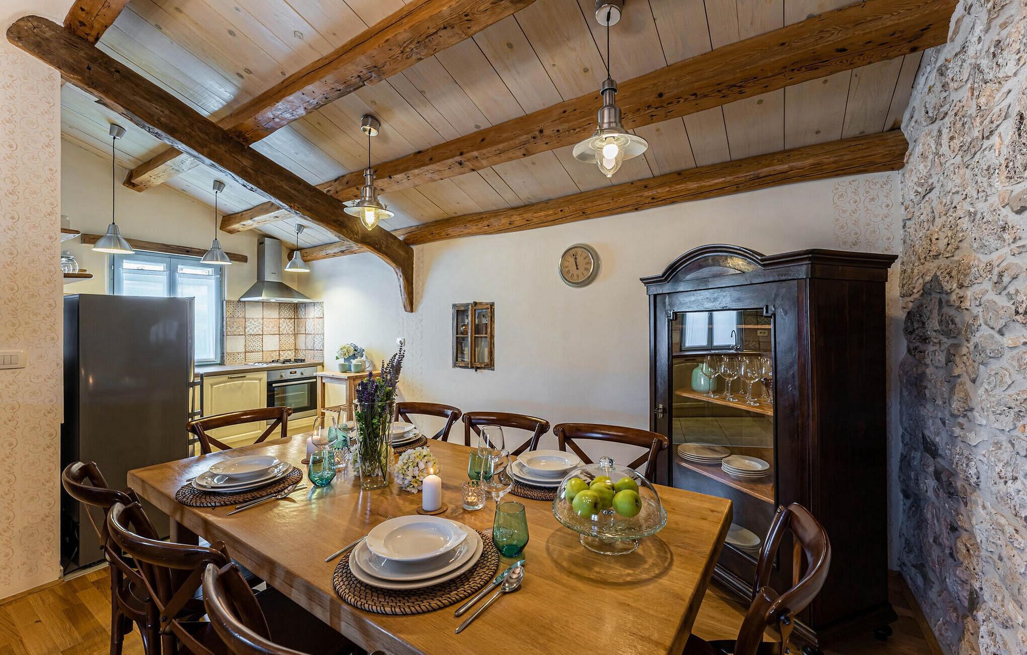 Un coin repas avec une table en bois, dressée pour quatre personnes, entourée de chaises. On aperçoit, au fond, une grande armoire en bois dotée de portes en verre.