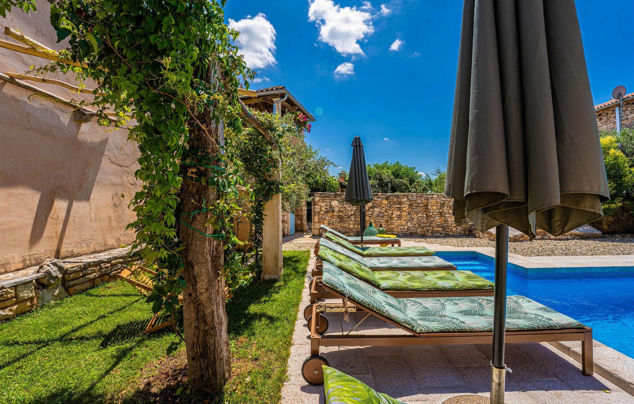 Zona de piscina con sillones de descanso y sombrillas, un muro de piedra y un cielo azul despejado.