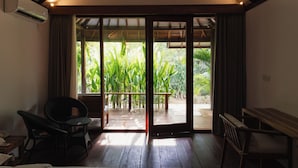 A room with a wooden floor, a sliding glass door leading to an outdoor area with plants, a chair, and a small table. - Villa Penyu (Gili Trawangan)