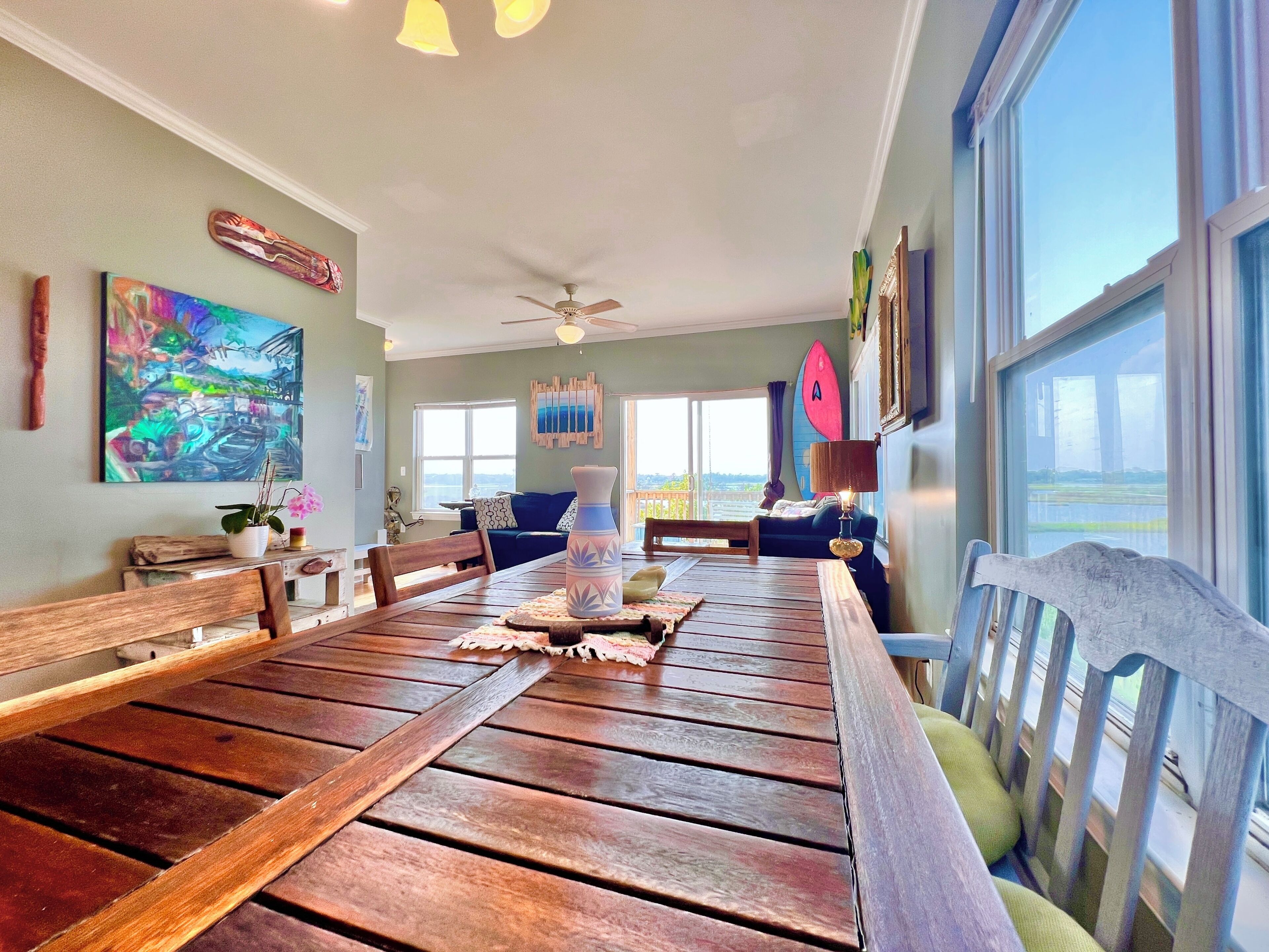 A dining area with a wooden table, chairs, and a ceiling fan. There are artworks on the walls and a view of the outdoors through large windows.