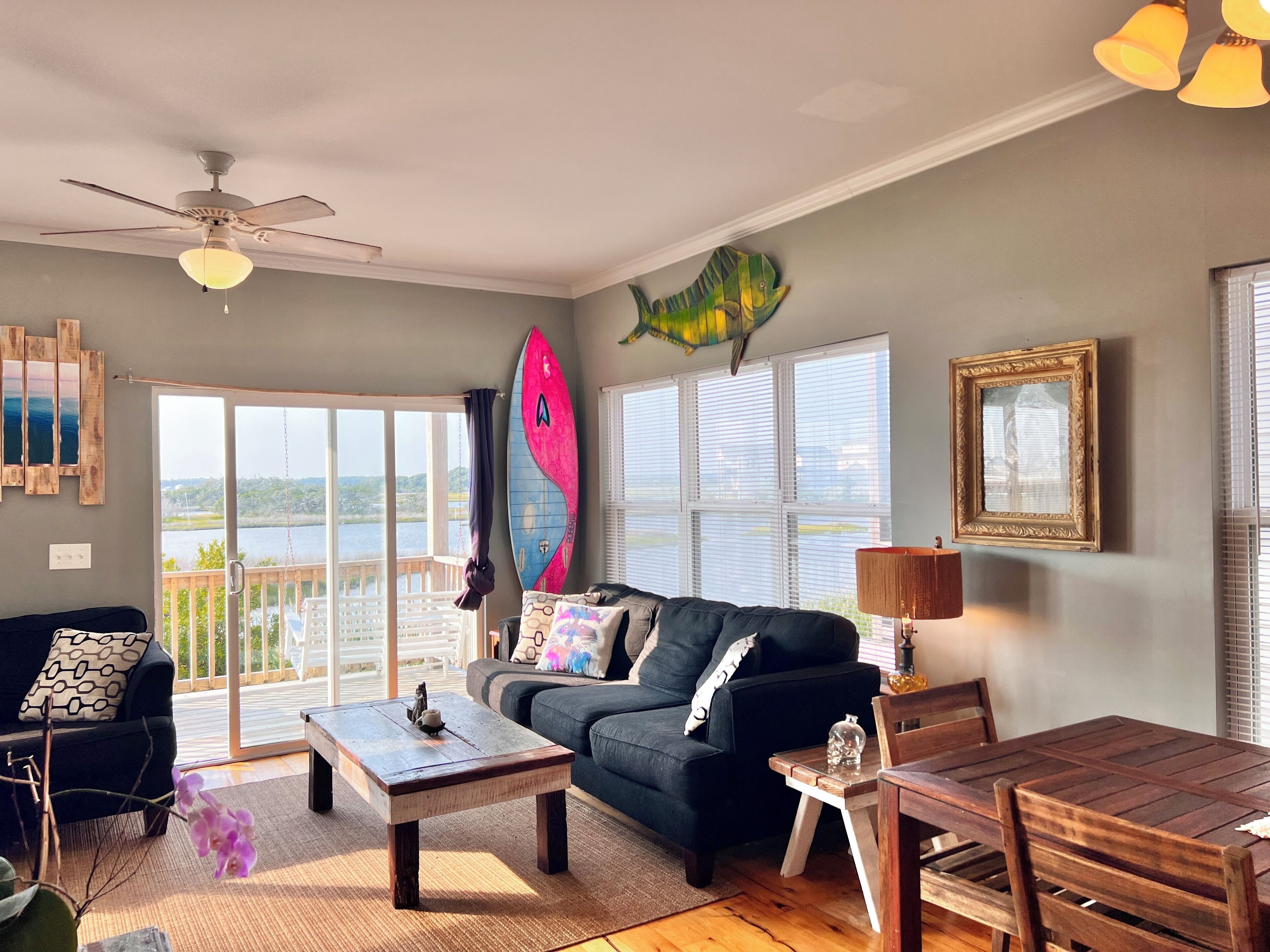 Una sala de estar con vista a un lago, un ventilador de techo, una tabla de surf y una mesa de comedor.