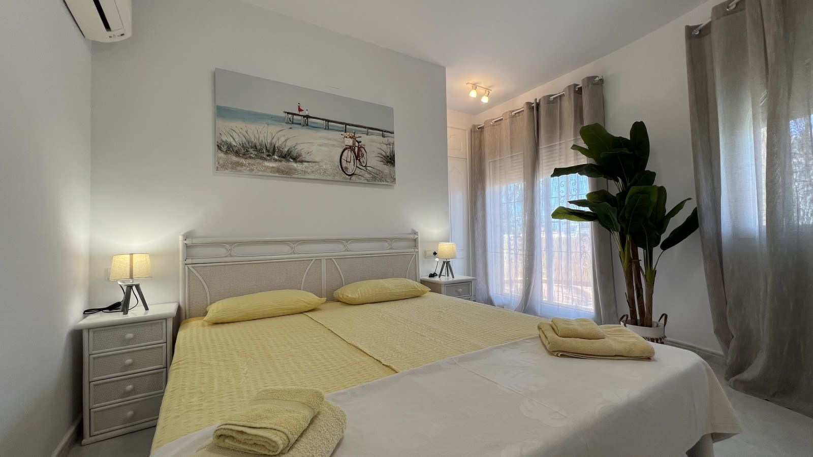 A hotel room with a bed, bedside tables, a lamp, a painting, a plant, and a window with curtains.