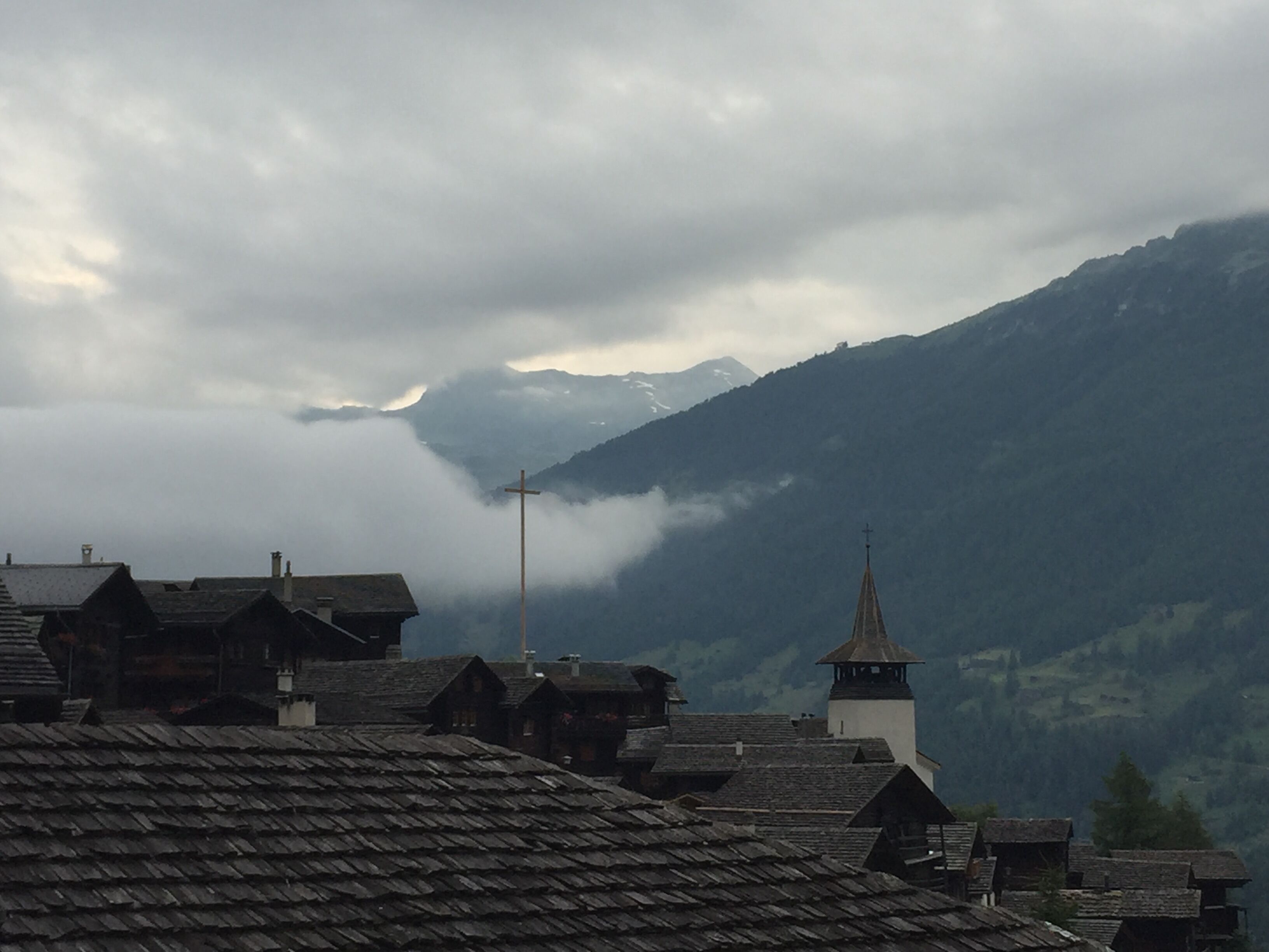 Een dorp met traditionele huizen en een kerk met een kruis, omringd door bergen en wolken.