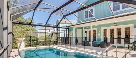 A covered swimming pool with a glass enclosure, adjacent to a building with a green exterior and a patio area with outdoor furniture.