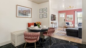 Een eethoek met een ronde tafel, roze bekleden stoelen en een bloemenvaas. Er hangen afbeeldingen in de muur en is uitzicht op een woonkamer met een bank en een televisie.