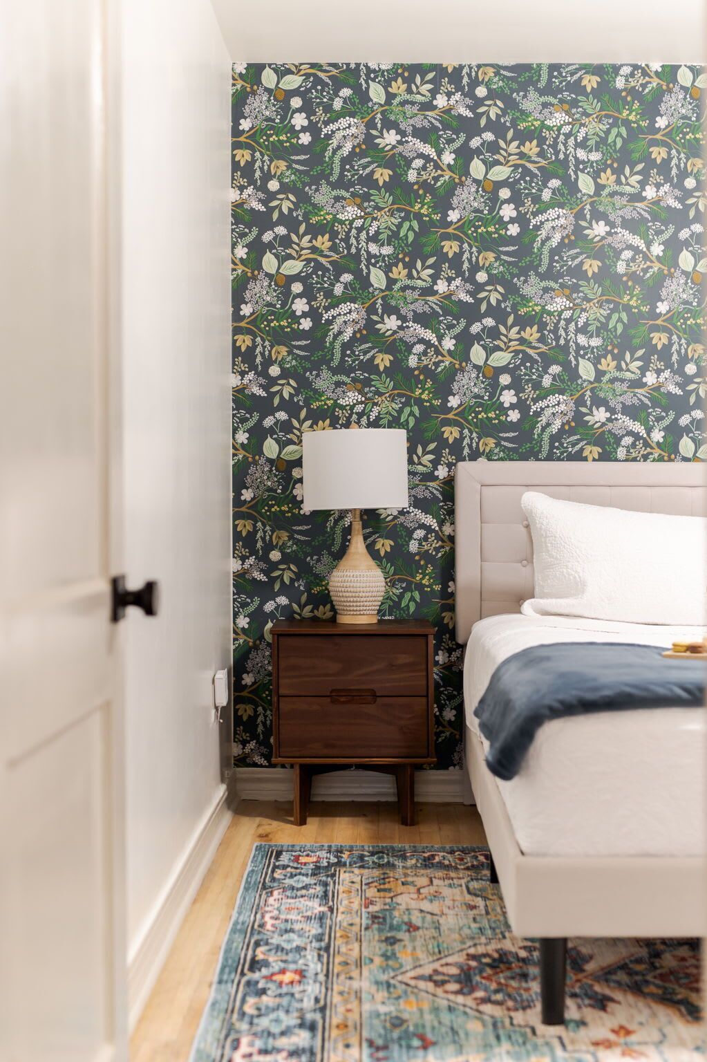 A bedroom with a floral wallpaper, a bedside table with a lamp, a bed with a white headboard, and a patterned rug.