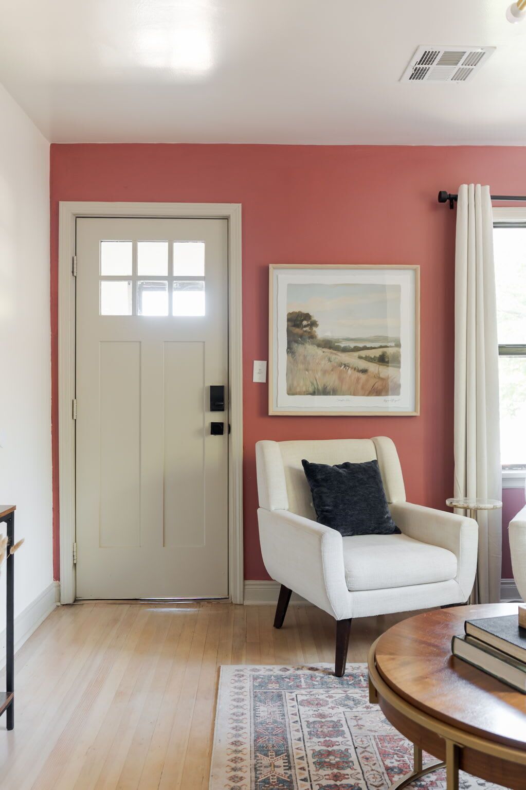 A room with a white door, a white armchair with a dark cushion, a framed landscape painting on the wall, a round wooden table with books, and a patterned rug on the floor.