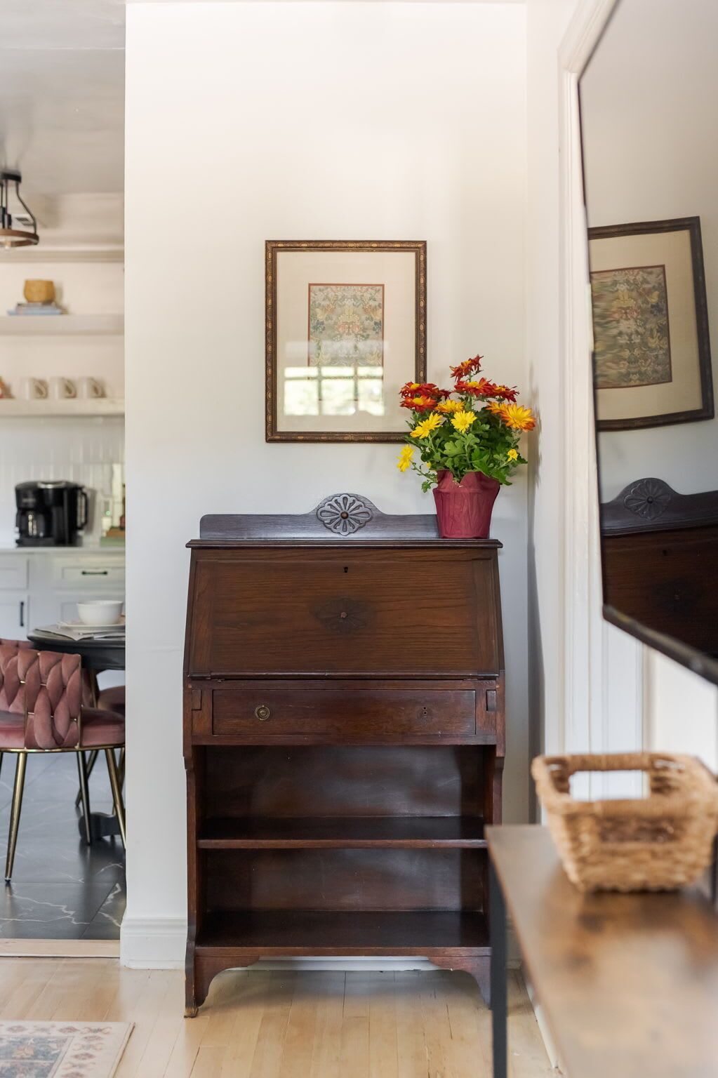 A wooden chest of drawers with a vase of flowers on top, a framed picture on the wall, and a wicker basket on a table.