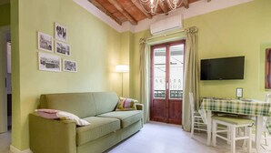 A living room with a green sofa, a dining table with chairs, a TV mounted on the wall, and a window with curtains. - 1BR Apt in Via Panicale by Sea N' Rent (Florence)