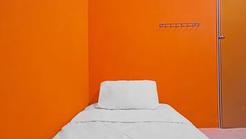 Un lit simple avec un matelas et un oreiller blancs, dans une pièce aux murs orange et dotée d’une porte.