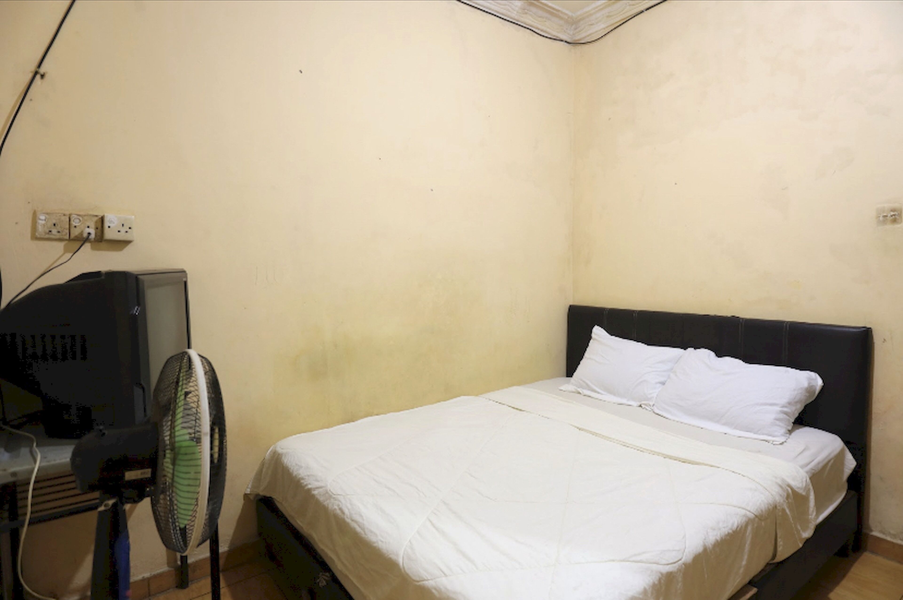 Una habitación pequeña con cama individual, una pared sencilla, un escritorio con computadora y un ventilador de pie.