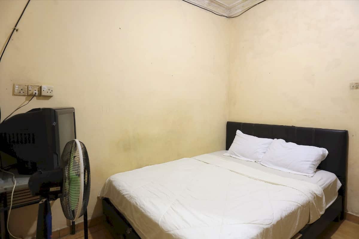Una habitación pequeña con cama individual, una pared sencilla, un escritorio con computadora y un ventilador de pie.
