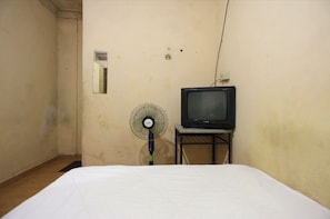 Una habitación pequeña y tenuemente iluminada, con una cama individual, un televisor sobre un pedestal y un ventilador de pie.