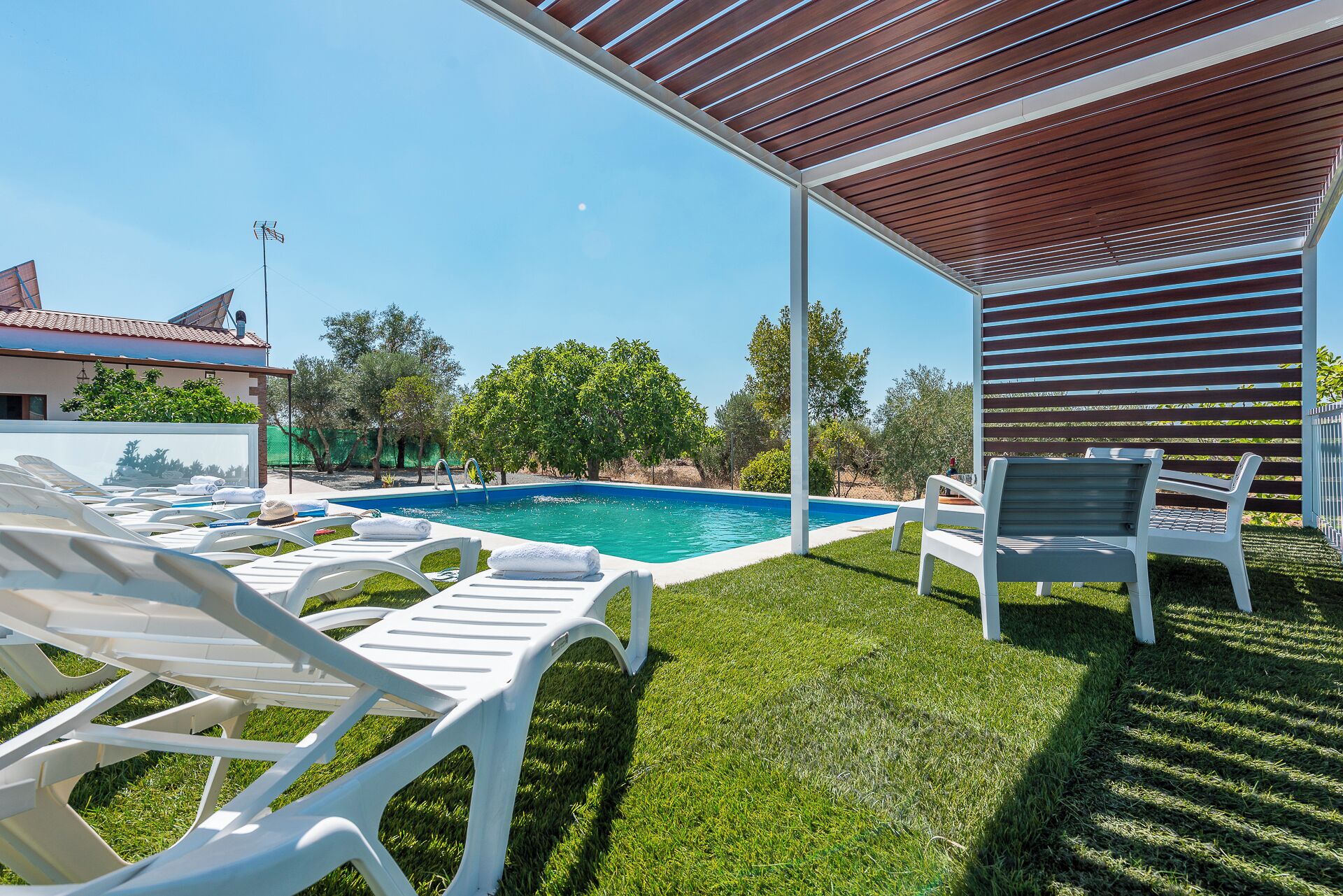 A poolside area with white lounge chairs, a swimming pool, and a covered patio with wooden slats.