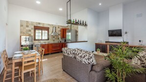 A modern kitchen with a built-in oven, microwave, and stainless steel refrigerator. There is a dining area with a wooden table and chairs, and a sofa with a floral throw. A flat-screen TV is mounted on the wall, and there is a small plant on the windowsill. - Holiday Home Adalid, Seville Countryside (La Puebla de Cazalla)