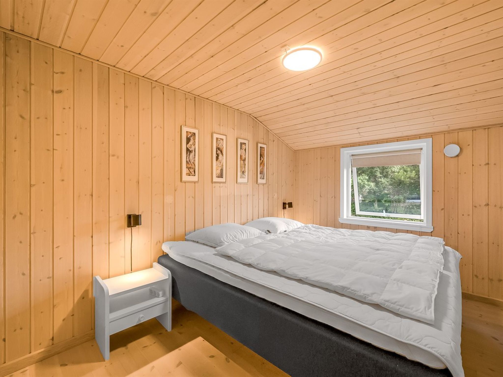 A bedroom with wooden walls, a bed with white bedding, a bedside table, and a window with a view of greenery.