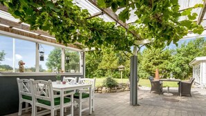 Un patio con mesa y sillas, una pérgola con parras y vistas al jardín circundante.