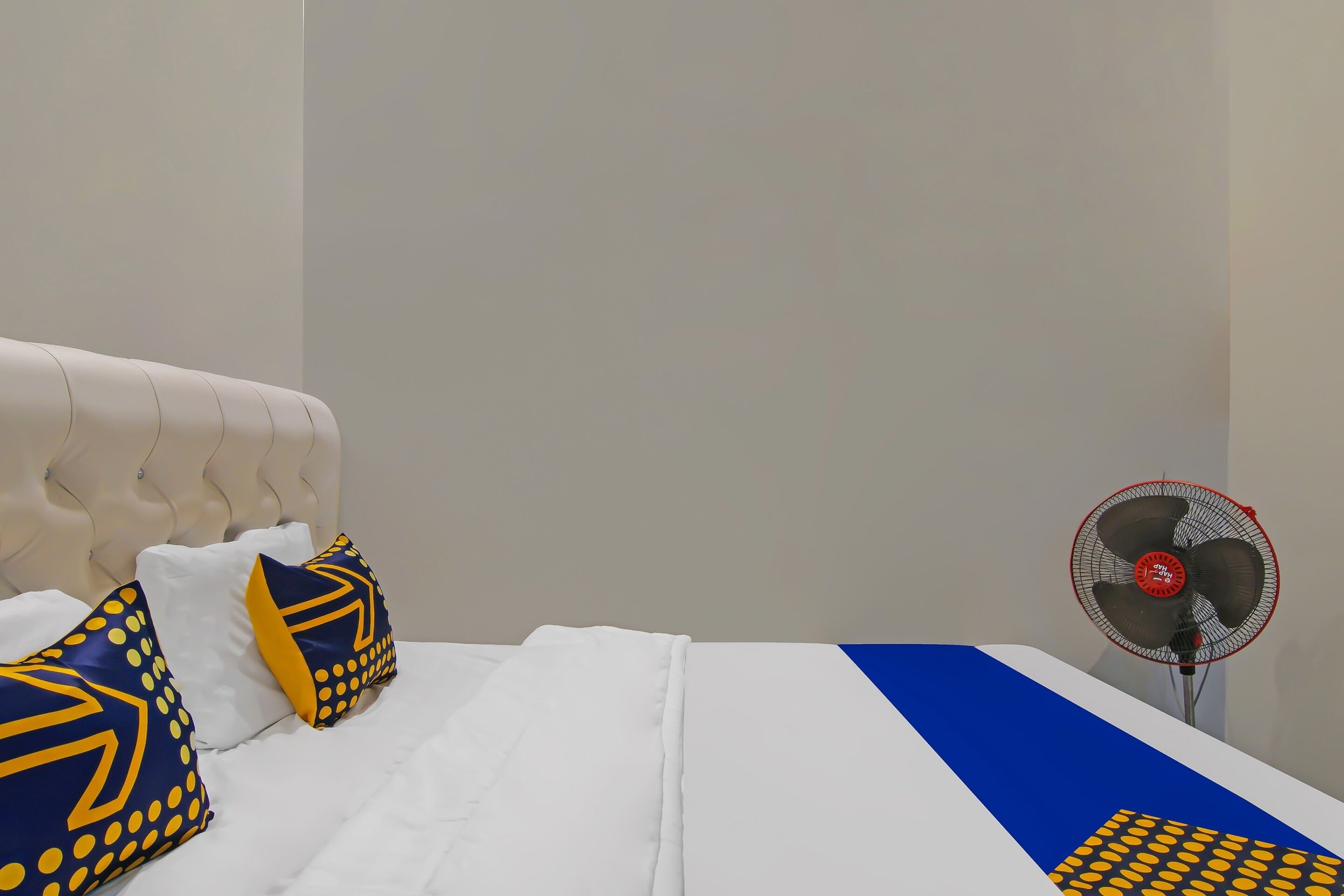 Cama com roupa de cama branca, almofadas azuis e amarelas, e um ventilador.