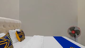 Cama com roupa de cama branca, almofadas azuis e amarelas, e um ventilador.