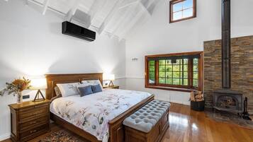 A bedroom with a wooden bed, a bench, a stone fireplace, and a window with a view of greenery.