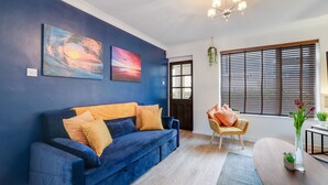 A living room with a blue sofa, a yellow armchair, and a wooden coffee table. There are two paintings on the wall, a door, and a window with blinds. - Bourne Court Attractive 1 bdrm Apartment (Ruislip)