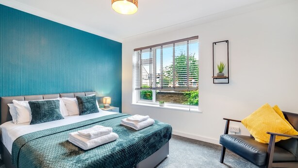 A modern bedroom with a large bed, a window with a view, a chair, and a bedside table. - Bourne Court Attractive 1 bdrm Apartment (Ruislip)