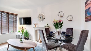A modern dining area with a glass table, cushioned chairs, a fireplace, and decorative wall art. - Bourne Court Attractive 1 bdrm Apartment (Ruislip)