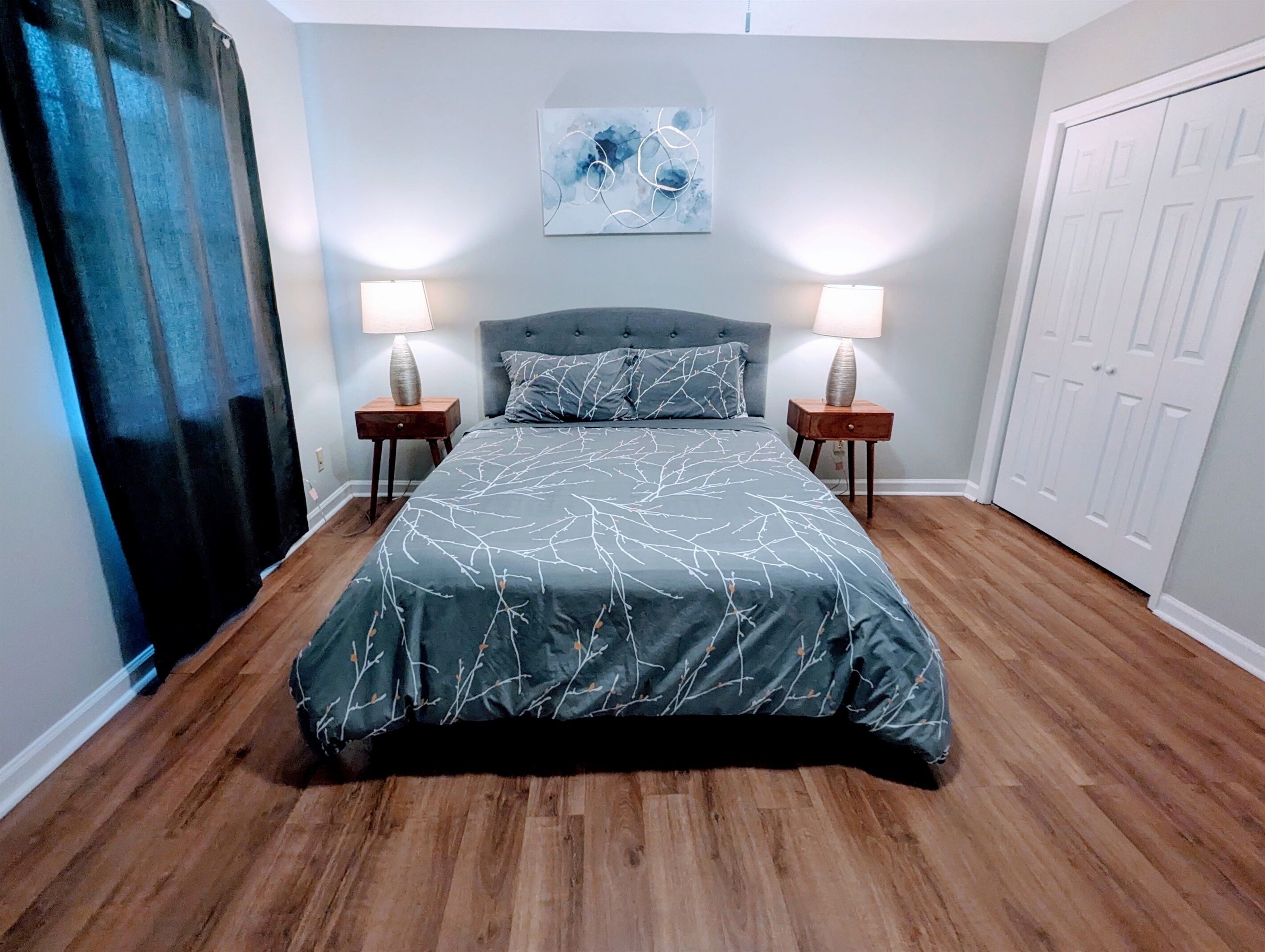 Une chambre à coucher avec un grand lit, deux tables de chevet, un tableau abstrait accroché au mur et une porte blanche.
