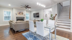 A modern living room with a fireplace, a dining area with a glass table, and a staircase. - Modern, Walkable Family Home w/ Porch & Parking (Atlanta)