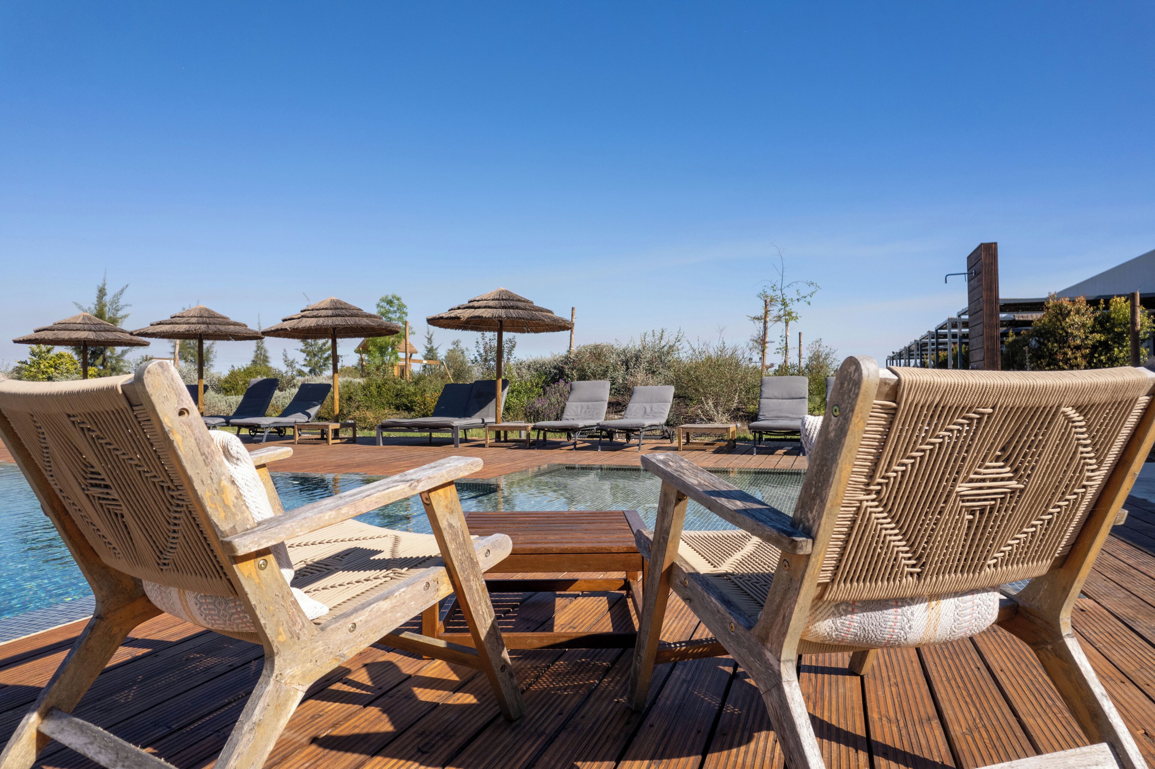 Eine Holzterrasse mit Liegestühlen und Sonnenschirmen, ein Schwimmbecken und ein Gebäude im Hintergrund.