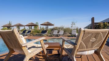 Eine Holzterrasse mit Liegestühlen und Sonnenschirmen, ein Schwimmbecken und ein Gebäude im Hintergrund.